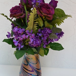 May include: A bouquet of purple roses and lilac flowers arranged in a swirling iridescent glass vase.