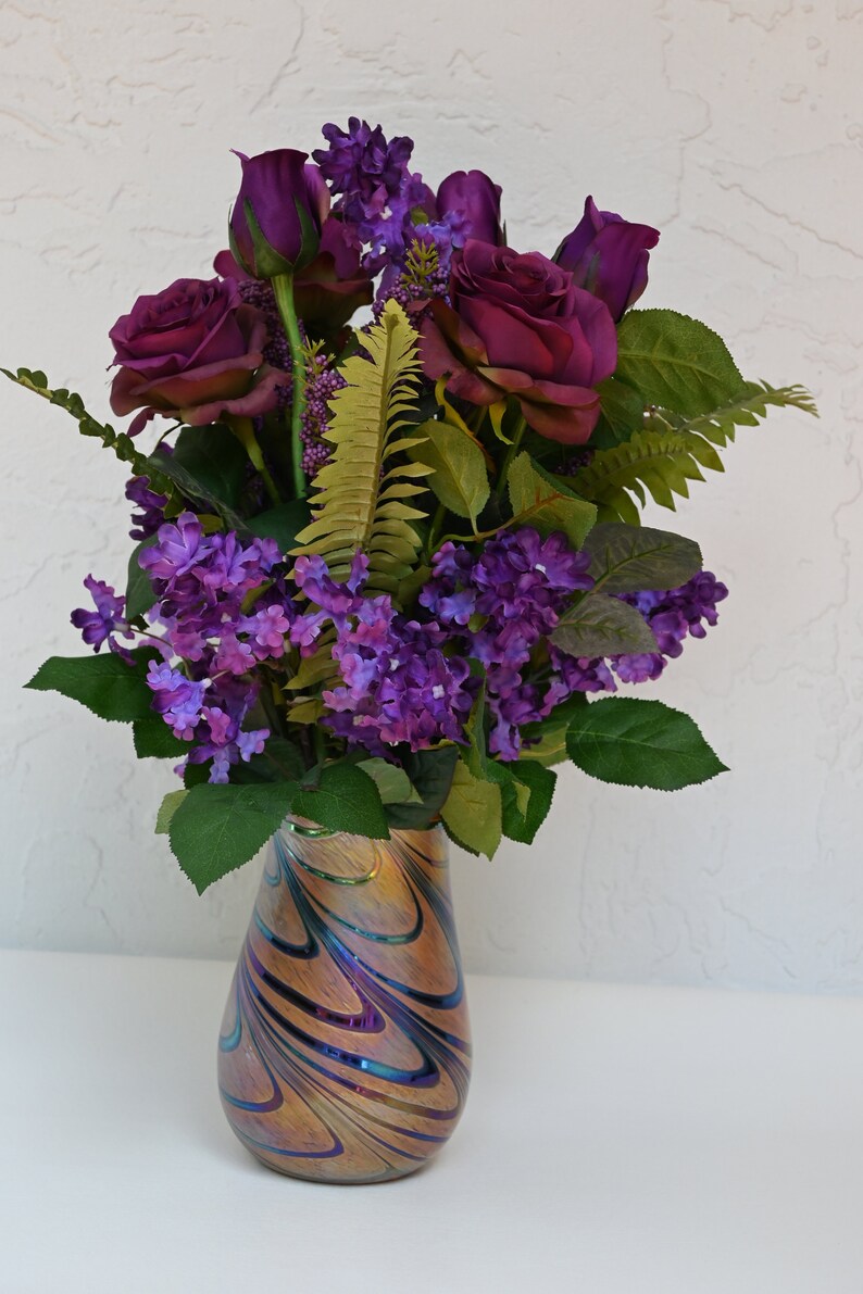 May include: A bouquet of purple roses and lilac flowers arranged in a swirling iridescent glass vase.