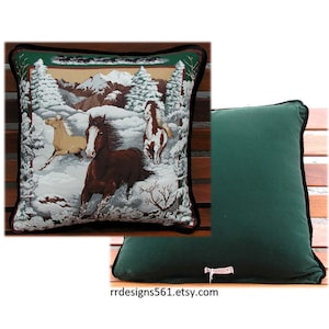 May include: A square throw pillow featuring a woven tapestry scene of three horses running through a snowy forest with mountains in the background. The reverse side of the pillow is solid dark green fabric with a small white tag at the bottom edge.