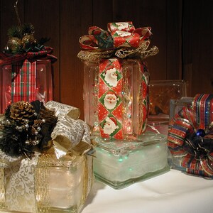 May include: Four square glass blocks decorated as holiday gifts. Each block is wrapped with patterned fabric ribbons and topped with bows, pinecones, or greenery. The glass blocks are illuminated from within by small, glowing string lights.