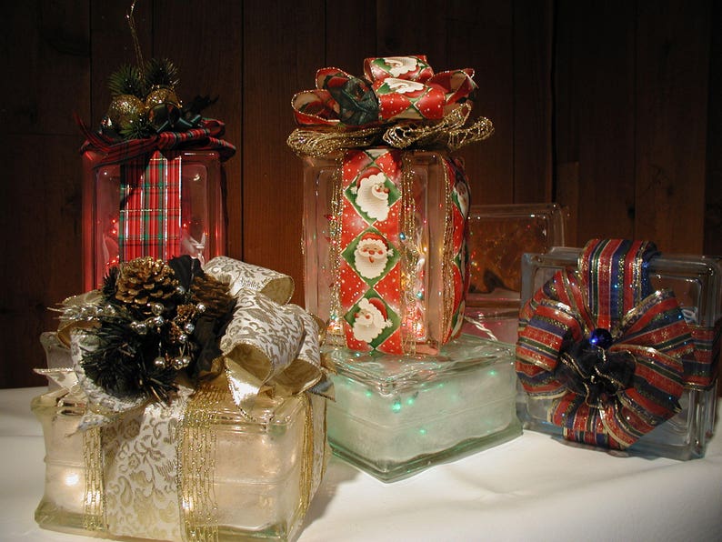 May include: Four square glass blocks decorated as holiday gifts. Each block is wrapped with patterned fabric ribbons and topped with bows, pinecones, or greenery. The glass blocks are illuminated from within by small, glowing string lights.