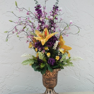 May include: A floral arrangement in a decorative bronze urn. The arrangement features yellow lilies, purple flowers, and delicate pink and white blossoms. The urn has a detailed relief design and sits on a square base, creating an elegant display.