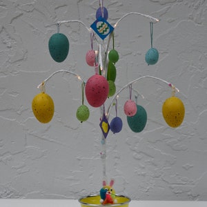 Op de afbeelding: Een witte metalen paasboom versierd met kleurrijke eieren en een konijnenfiguur in een gele pot. De boom is versierd met een blauw en groen ruitvormig ornament.