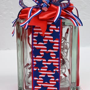 May include: A rectangular glass block filled with white string lights featuring red bulbs. The block is wrapped in a vertical ribbon with a blue star and red and white stripe pattern, topped with a multi-layered bow of matching ribbon and a small star-shaped charm.