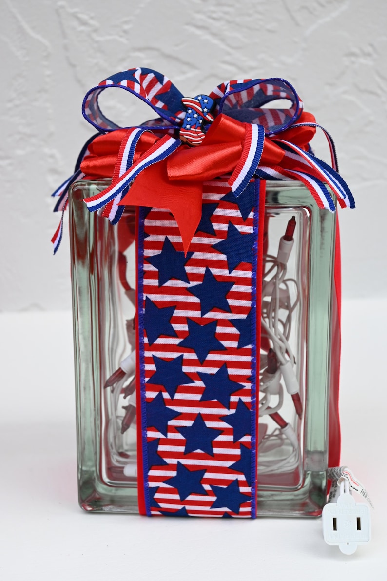 May include: A rectangular glass block filled with white string lights featuring red bulbs. The block is wrapped in a vertical ribbon with a blue star and red and white stripe pattern, topped with a multi-layered bow of matching ribbon and a small star-shaped charm.