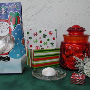 Op de afbeelding: Een rood-wit gestreepte geschenkdoos staat bovenop een groen-wit gepatroneerde geschenkdoos. Een rode glazen pot met deksel staat op een groene ondergrond. Een witte koek ligt op een doorzichtig bord. Een witte dennenappel ligt op de groene ondergrond. De afbeelding is versierd met een witte achtergrond met sneeuwvlokken.