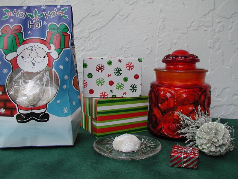 Op de afbeelding: Een rood-wit gestreepte geschenkdoos staat bovenop een groen-wit gepatroneerde geschenkdoos. Een rode glazen pot met deksel staat op een groene ondergrond. Een witte koek ligt op een doorzichtig bord. Een witte dennenappel ligt op de groene ondergrond. De afbeelding is versierd met een witte achtergrond met sneeuwvlokken.
