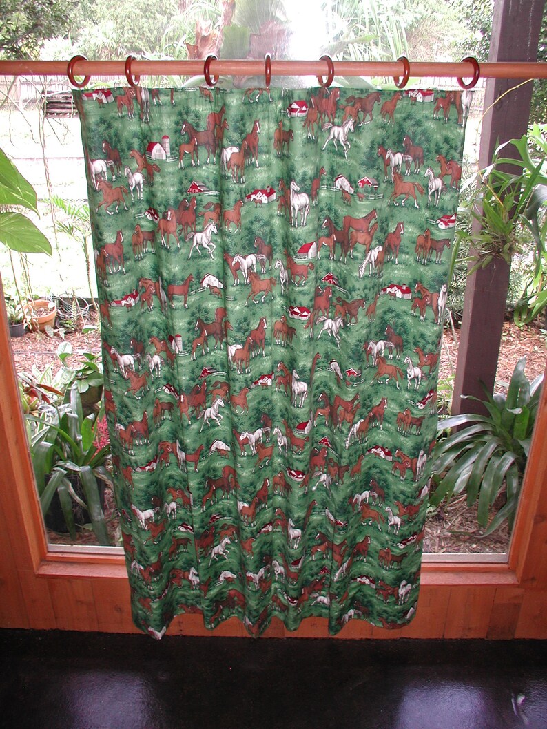 May include: A green fabric curtain featuring a repeating pattern of brown, white, and black horses in a field with small red barns. The curtain is suspended from a horizontal wooden rod by several circular brown rings along the top edge.