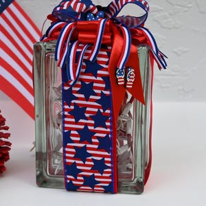 May include: A rectangular glass block filled with white string lights, wrapped in a decorative ribbon featuring blue stars and red and white stripes. The top is adorned with a multi-layered bow in red, white, and blue, accented with small flip-flop charms.