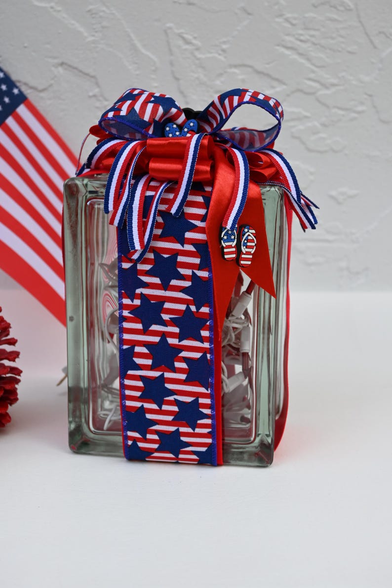 May include: A rectangular glass block filled with white string lights, wrapped in a decorative ribbon featuring blue stars and red and white stripes. The top is adorned with a multi-layered bow in red, white, and blue, accented with small flip-flop charms.
