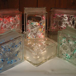 May include: A collection of five rectangular glass blocks, each containing a string of miniature electric lights. The lights inside the blocks glow in various colors, including red, blue, green, and multi-colored, with white wiring visible.