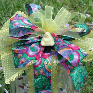 May include: A colorful ribbon bow with a green, pink, and purple pattern, tied around a clear glass jar. The bow is decorated with a yellow chick figurine and a green and white polka dot ribbon.