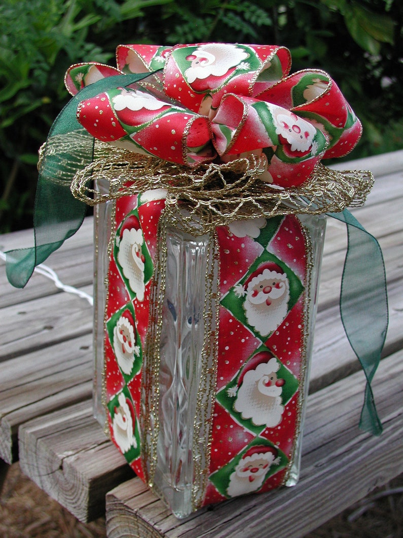 May include: A square glass block wrapped in red and green ribbon featuring a repeating Santa Claus pattern. The block is topped with a large, multi-layered bow made of the same Santa ribbon, accented with a sheer gold mesh ribbon and green trim.