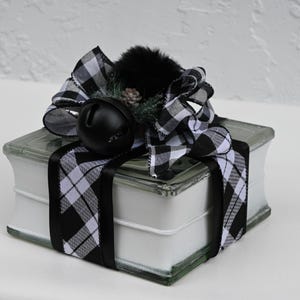 May include: A square glass block with white interior, wrapped in black and white buffalo plaid ribbon. The top is decorated with a large black jingle bell, a black faux fur pom-pom, and a small artificial pine sprig with a tiny pinecone.