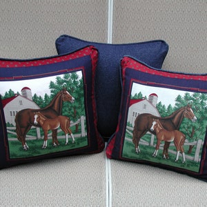 May include: Two square throw pillows featuring a central graphic of a brown mare and foal in a green pasture with a white barn and silo. The pillows have a dark blue border and a red patterned outer edge. A third solid dark blue pillow sits behind.