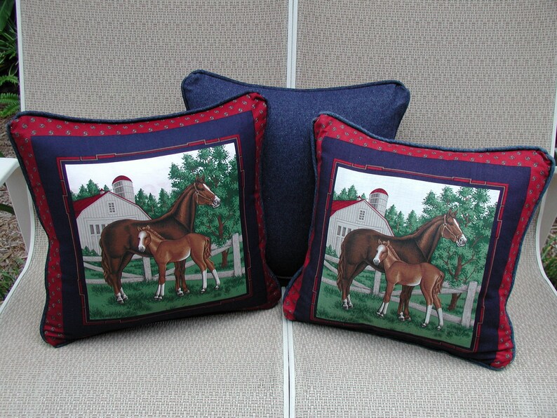 May include: Two square throw pillows featuring a central graphic of a brown mare and foal in a green pasture with a white barn and silo. The pillows have a dark blue border and a red patterned outer edge. A third solid dark blue pillow sits behind.