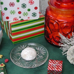 Op de afbeelding: Een witte gepoederde koek op een helder glazen bord met een rood-wit gestreepte geschenkdoos, een rode glazen pot met deksel en een groen-wit gestreepte geschenkdoos op de achtergrond.