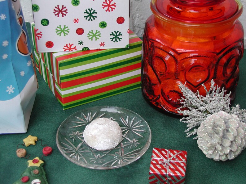 Op de afbeelding: Een witte gepoederde koek op een helder glazen bord met een rood-wit gestreepte geschenkdoos, een rode glazen pot met deksel en een groen-wit gestreepte geschenkdoos op de achtergrond.