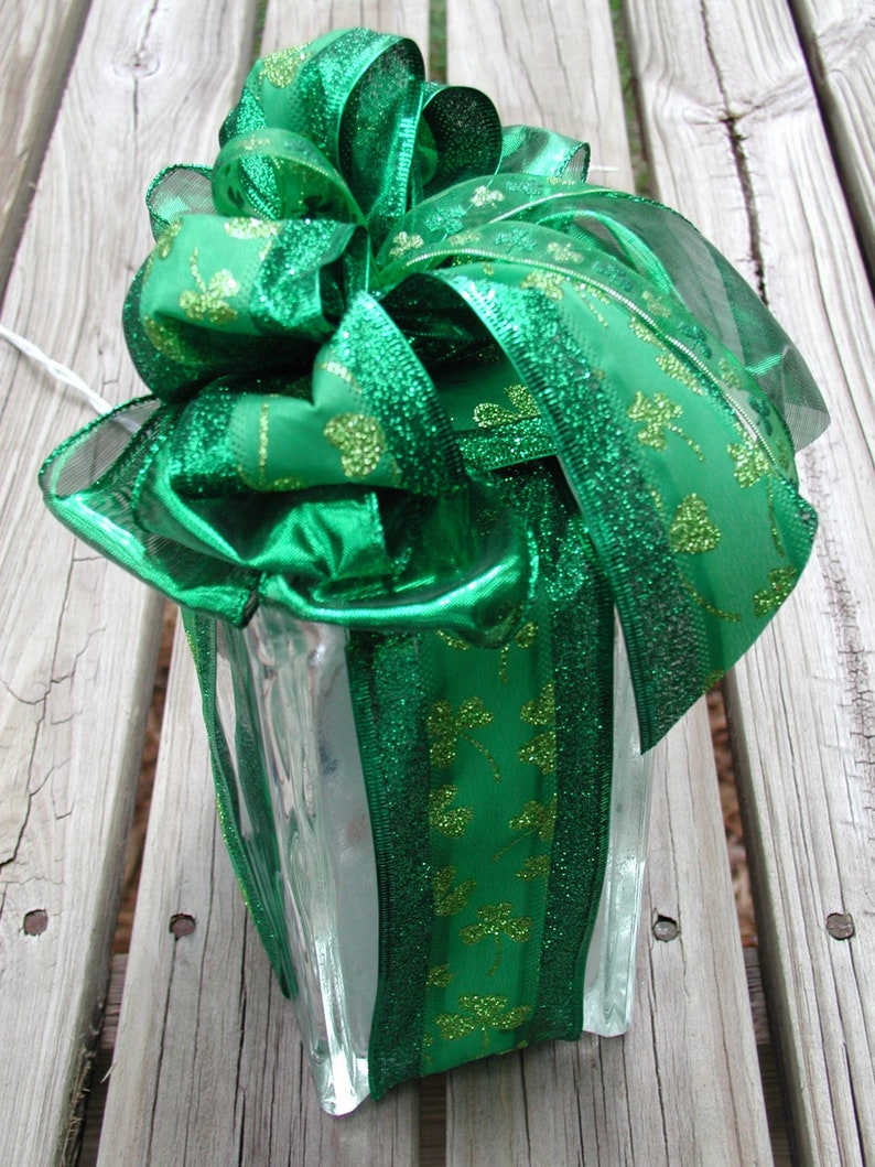 May include: A clear glass block with a textured surface, topped with a large, multi-looped green ribbon. The ribbon features a repeating pattern of glittery green shamrocks and has shimmering edges, creating a decorative accent piece.