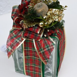May include: A square glass block wrapped in red and green plaid ribbon with gold trim. The top is decorated with a large bow, a textured gold sphere, small gold ornaments, and artificial pine needles. A white electrical cord extends from the side.