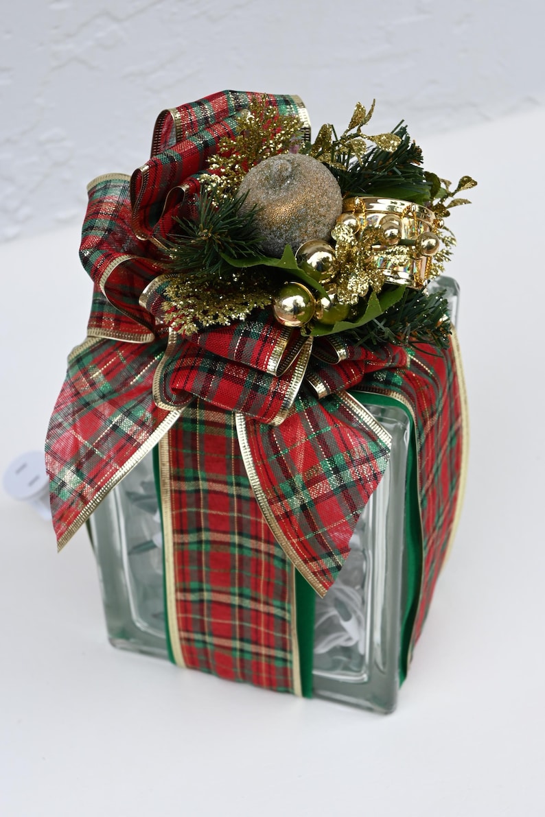 May include: A square glass block wrapped in red and green plaid ribbon with gold trim. The top is decorated with a large bow, a textured gold sphere, small gold ornaments, and artificial pine needles. A white electrical cord extends from the side.