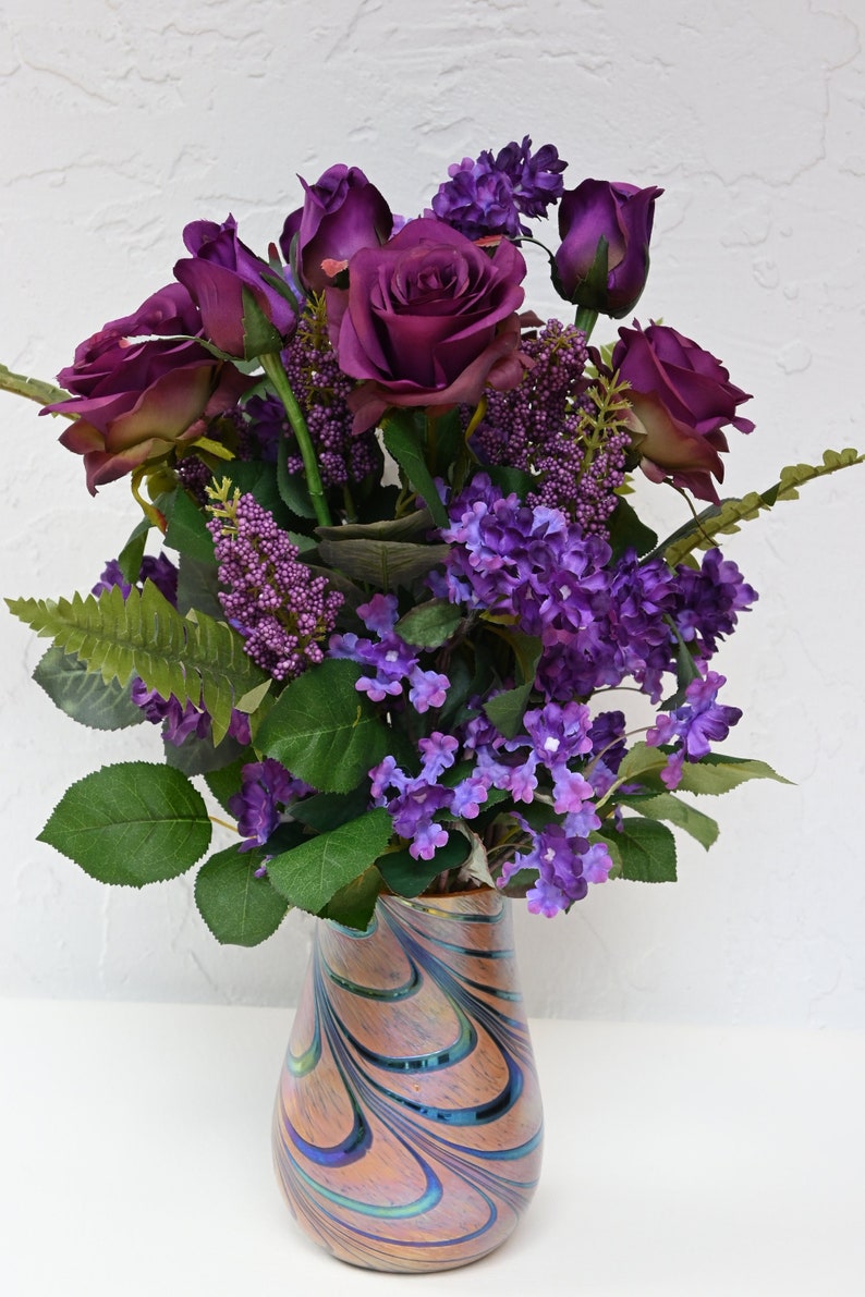 May include: A bouquet of purple roses and lilac flowers arranged in a swirling iridescent glass vase.