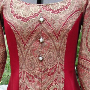May include: A red dress with a gold and red paisley pattern. The dress has a scoop neckline and long sleeves. The dress is decorated with four white buttons.