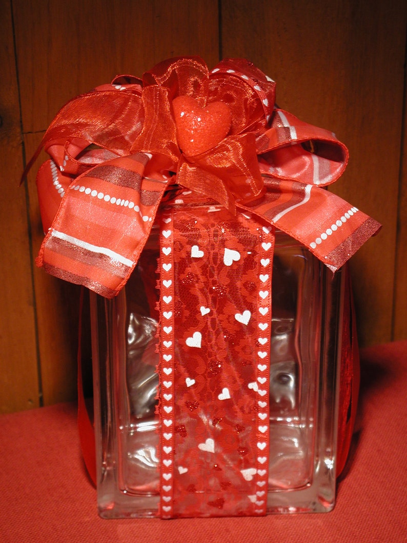 May include: A clear glass block wrapped in a red lace ribbon featuring small white heart patterns. The top is adorned with a large, multi-layered red fabric bow and a single, central, glittery red heart ornament.