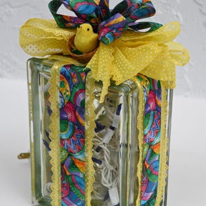 May include: A rectangular glass block filled with white string lights, wrapped in colorful patterned ribbon featuring Easter eggs. The top is decorated with a large yellow polka-dot bow and a small yellow felt chick sitting in a tiny nest.