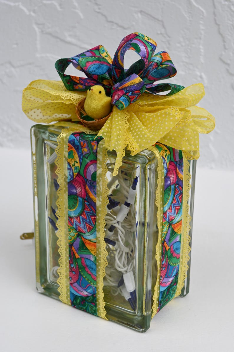 May include: A rectangular glass block filled with white string lights, wrapped in colorful patterned ribbon featuring Easter eggs. The top is decorated with a large yellow polka-dot bow and a small yellow felt chick sitting in a tiny nest.