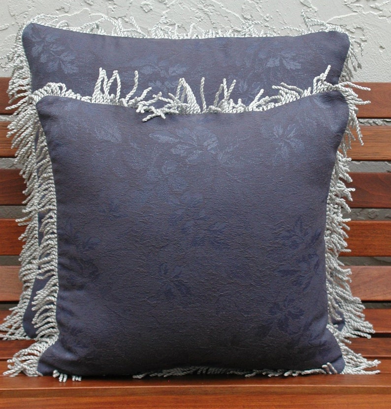 May include: Two decorative throw pillows with a dark blue fabric and a white floral pattern. The pillows have a white fringe trim.