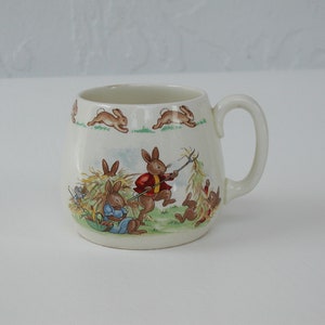 May include: Cream-colored ceramic mug with a curved handle. The mug features a pastoral scene with illustrated rabbits in various activities, including running and gathering hay. The top of the mug has a repeating pattern of running rabbits.