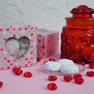 Op de afbeelding: Twee roze dozen met hartvormige uitsparingen gevuld met witte koekjes. Een rode glazen pot met een deksel en een rood hartontwerp staat op een roze oppervlak met rode snoephartjes die eromheen liggen.