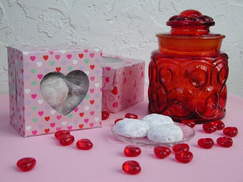 Op de afbeelding: Twee roze dozen met hartvormige uitsparingen gevuld met witte koekjes. Een rode glazen pot met een deksel en een rood hartontwerp staat op een roze oppervlak met rode snoephartjes die eromheen liggen.