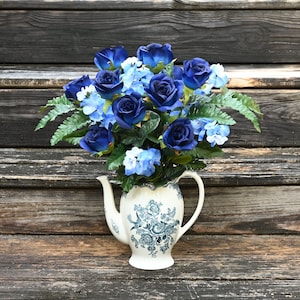 Puede incluir: Una tetera azul y blanca con un diseño floral contiene un ramo de rosas azules e hortensias azules. Las flores están dispuestas en un estilo vintage y se encuentran sobre un fondo de madera rústica.