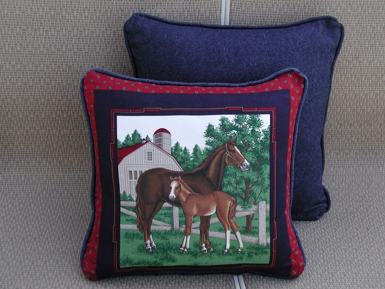 May include: A square decorative pillow featuring a central panel with a brown horse and foal standing in a green pasture before a white barn and silo. The panel is framed by a dark blue border and a red outer border with a small repeating pattern.