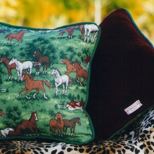 May include: Two square throw pillows with green piping. One pillow features a green fabric with a repeating pattern of brown, white, and black horses in a field with red barns. The other pillow is made of solid dark maroon velvet fabric.