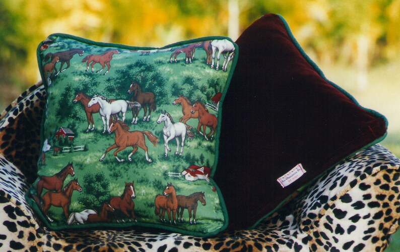 May include: Two square throw pillows with green piping. One pillow features a green fabric with a repeating pattern of brown, white, and black horses in a field with red barns. The other pillow is made of solid dark maroon velvet fabric.