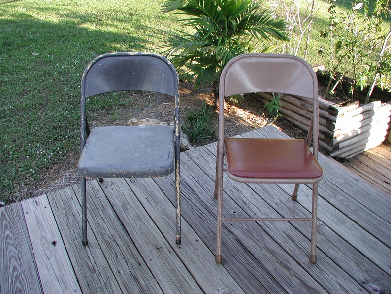 May include: Two folding chairs, one with a gray metal seat and back, the other with a brown vinyl seat and a beige metal frame. Both chairs are on a wooden deck.