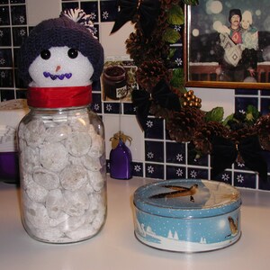 Op de afbeelding: Een glazen pot gevuld met witte poedersuikerkoekjes, bedekt met een sneeuwpopfiguur met een blauwe gebreide muts en een rode sjaal. De pot staat op een wit aanrecht met een blauwe en witte betegelde achterwand.