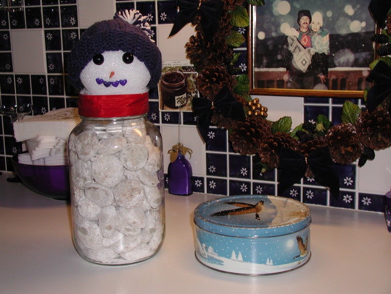 Op de afbeelding: Een glazen pot gevuld met witte poedersuikerkoekjes, bedekt met een sneeuwpopfiguur met een blauwe gebreide muts en een rode sjaal. De pot staat op een wit aanrecht met een blauwe en witte betegelde achterwand.
