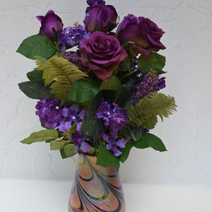 May include: A bouquet of purple roses and small purple flowers in a swirled glass vase with iridescent colors.