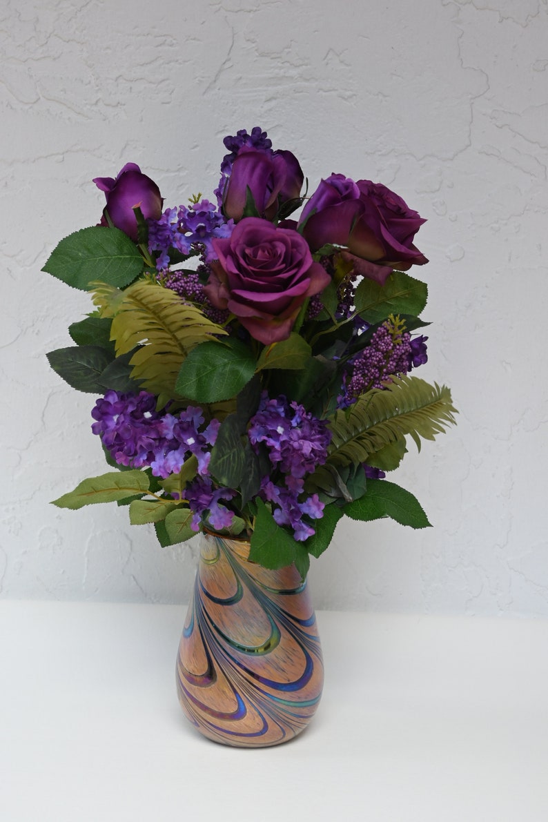 May include: A bouquet of purple roses and small purple flowers in a swirled glass vase with iridescent colors.