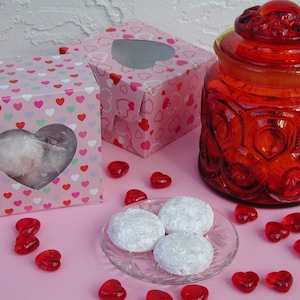 Op de afbeelding: Drie hartvormige koekjes bestoven met poedersuiker op een heldere glazen plaat met rode hartvormige snoepjes die eromheen zijn verspreid. Twee roze dozen met hartvormige uitsnijdingen en een rode glazen pot met deksel staan op de achtergrond.