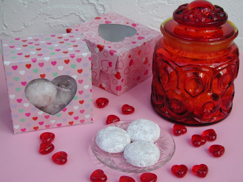 Op de afbeelding: Drie hartvormige koekjes bestoven met poedersuiker op een heldere glazen plaat met rode hartvormige snoepjes die eromheen zijn verspreid. Twee roze dozen met hartvormige uitsnijdingen en een rode glazen pot met deksel staan op de achtergrond.