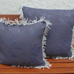 May include: Two decorative throw pillows with a blue floral pattern and white fringe trim. The pillows are on a wooden bench.