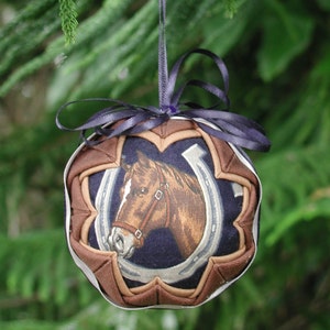 May include: A brown and white quilted Christmas ornament with a picture of a brown horse and a horseshoe on a navy blue background. The ornament has a purple ribbon.