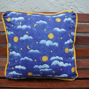 May include: A blue pillow with a yellow border features a repeating pattern of white clouds, yellow suns, and silver moons on a blue background.