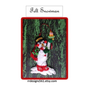 May include: A white felt snowman ornament featuring a black top hat, a red and green scarf, and red boots. The snowman holds a small bell with the text NOEL printed on it. The ornament is decorated with sequins and includes a red hanging ribbon.