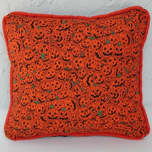 K&ouml;nnte beinhalten: Orangefarbener Kissenbezug mit Halloween-Motiv, das ein sich wiederholendes Muster aus schwarzen K&uuml;rbisgesichtern zeigt.